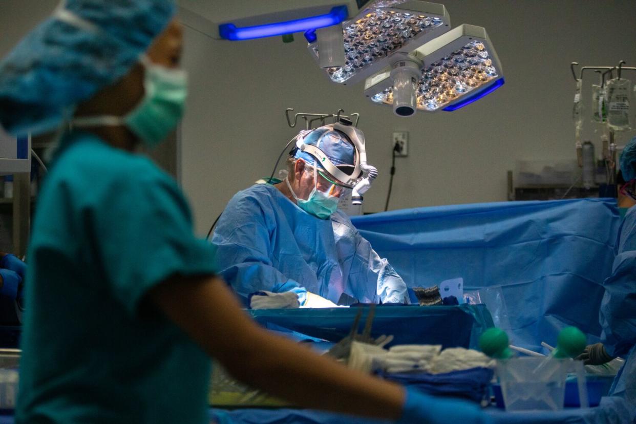 A doctor, wearing scrubs, mask, face shield and medical head gear, stands behind a covered operating table.