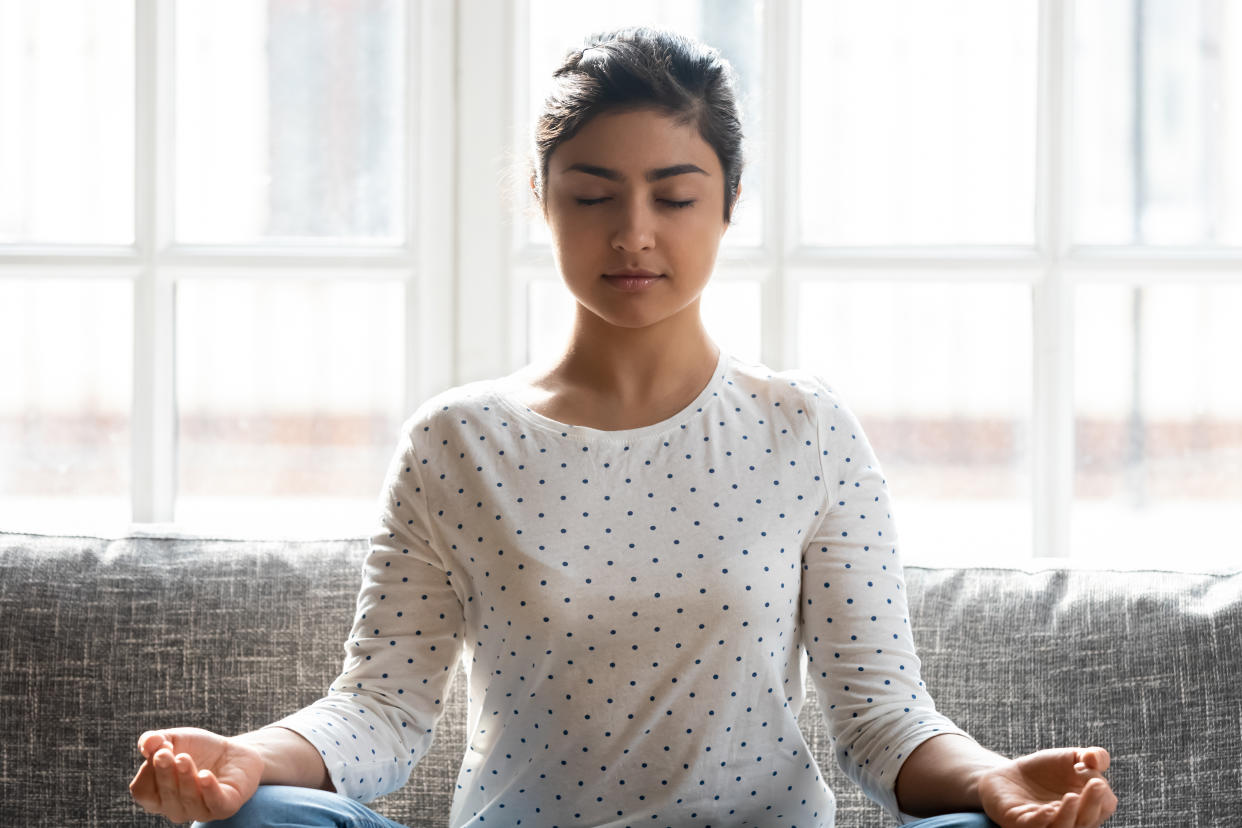 Calm Indian woman with closed eyes meditating at home alone close up, relaxed beautiful girl sitting in lotus pose on comfortable couch, dreaming, doing yoga exercise, stress relief concept