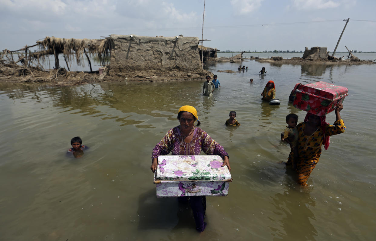 One woman carries a large box in her arms, while another balances one on her head, as she holds a child in one arm, as they make their way through waist-deep floodwaters from a small hut. Children swim in the water beside them.