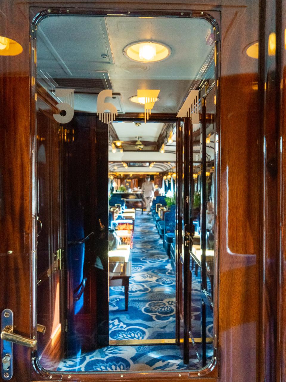 A shiny, windowed, wooden train door leads to a carriage with blue carpets and seating.