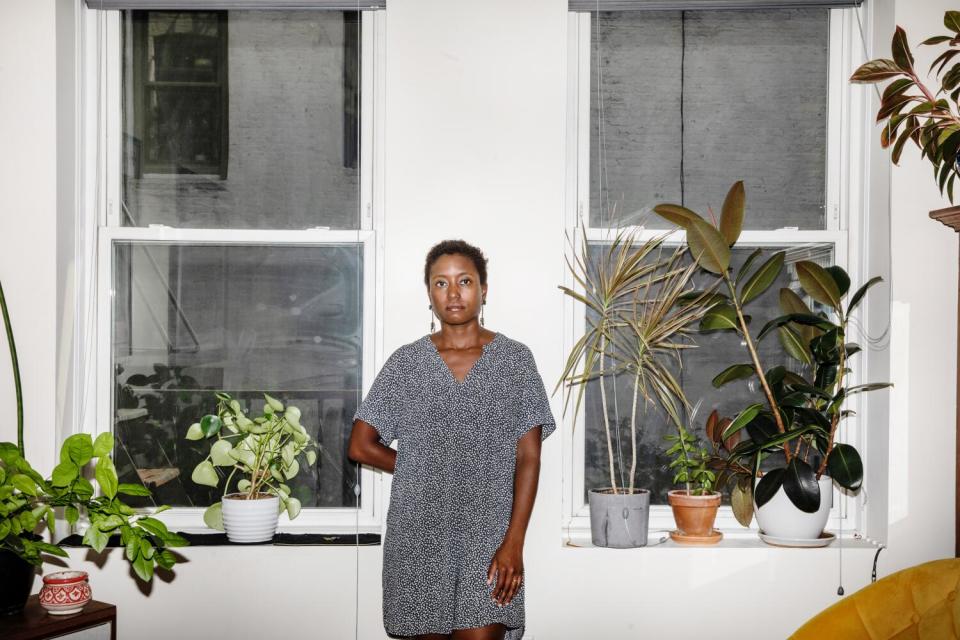 A woman poses in front of a wall with a pair of windows, surrounded by houseplants.
