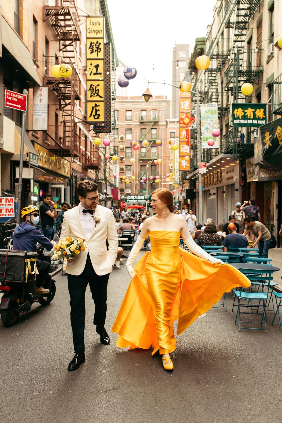A bride in a yellow dress and a groom in a white jacket and black pants walk through China town.
