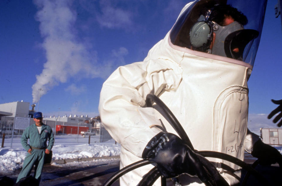 Person in a protective suit handling equipment with another person observing in a snowy environment