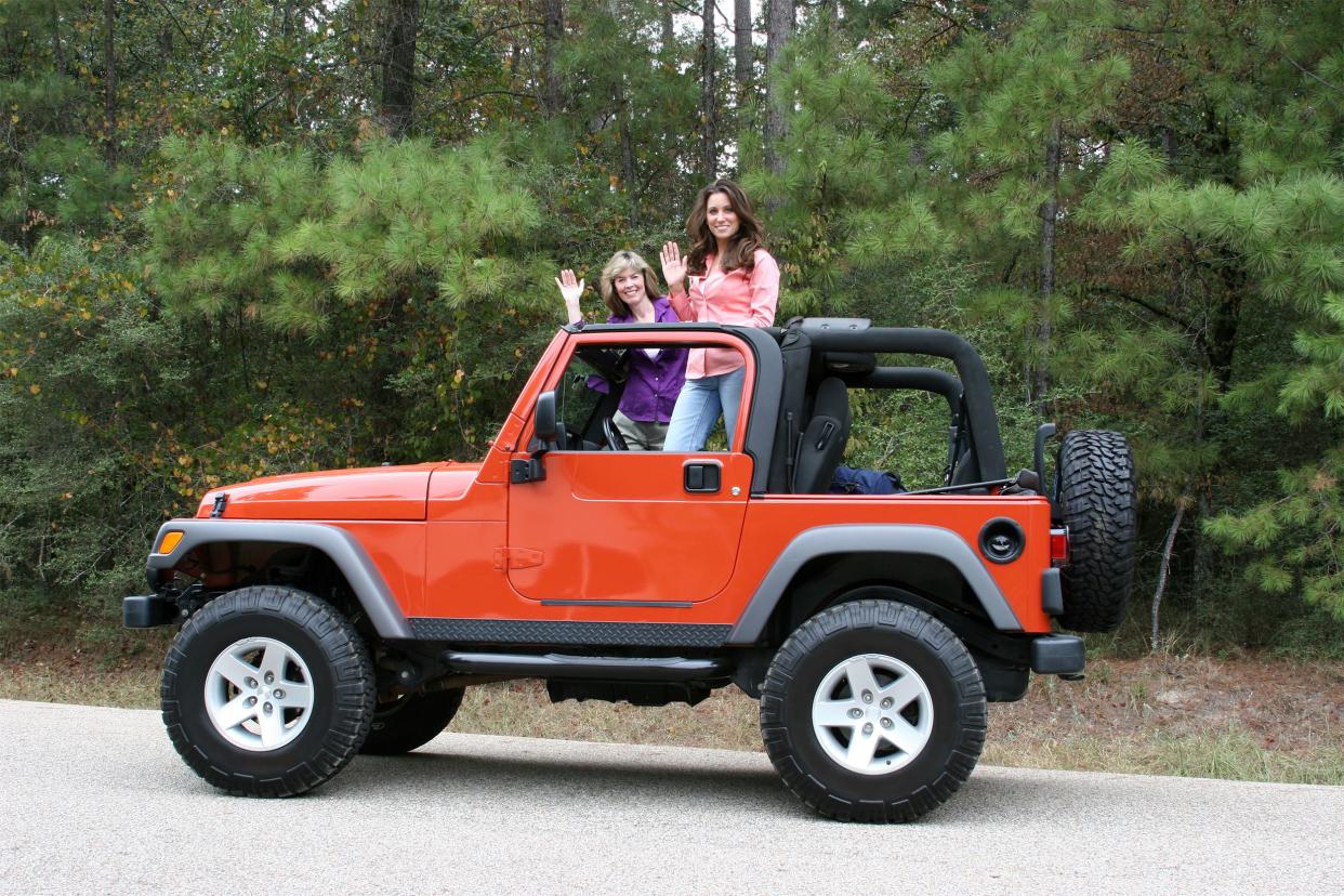 Two women standing and waving in a small orange Jeep.