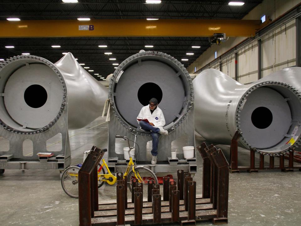 worker sits inside the base of a giant tube shaped wind turbine blade on its side inside manufacturing garage facility