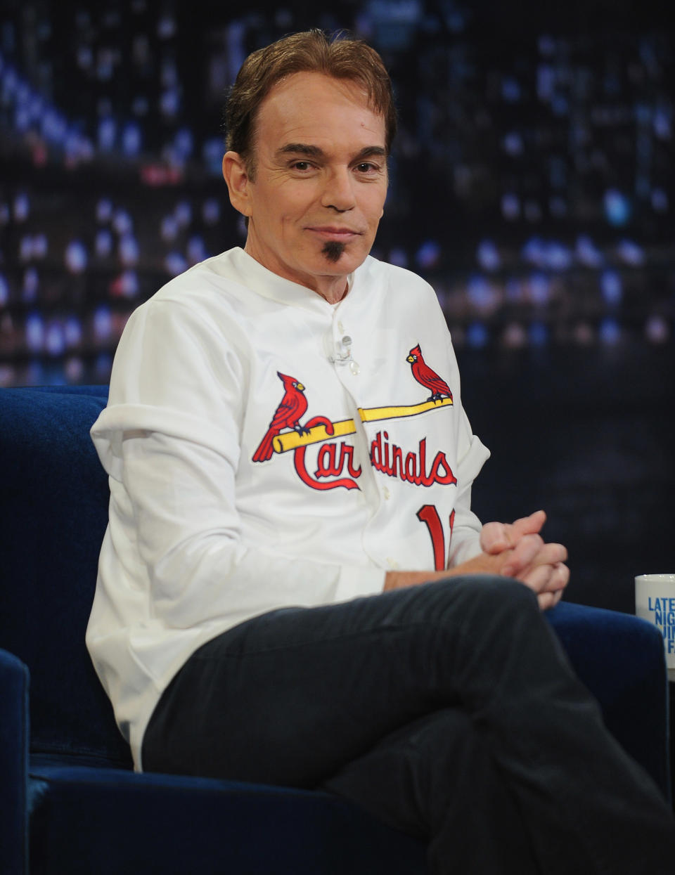 Billy Bob Thornton, wearing a St. Louis Cardinals baseball jersey, sits cross-legged on a talk show set with a cityscape background