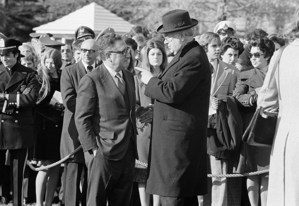 Soviet Ambassador Anatoly Dobrynin, in bowler hat, points his index finger at Henry Kissinger, who has his hands in his pockets, with a crowd of spectators behind a rope line.  