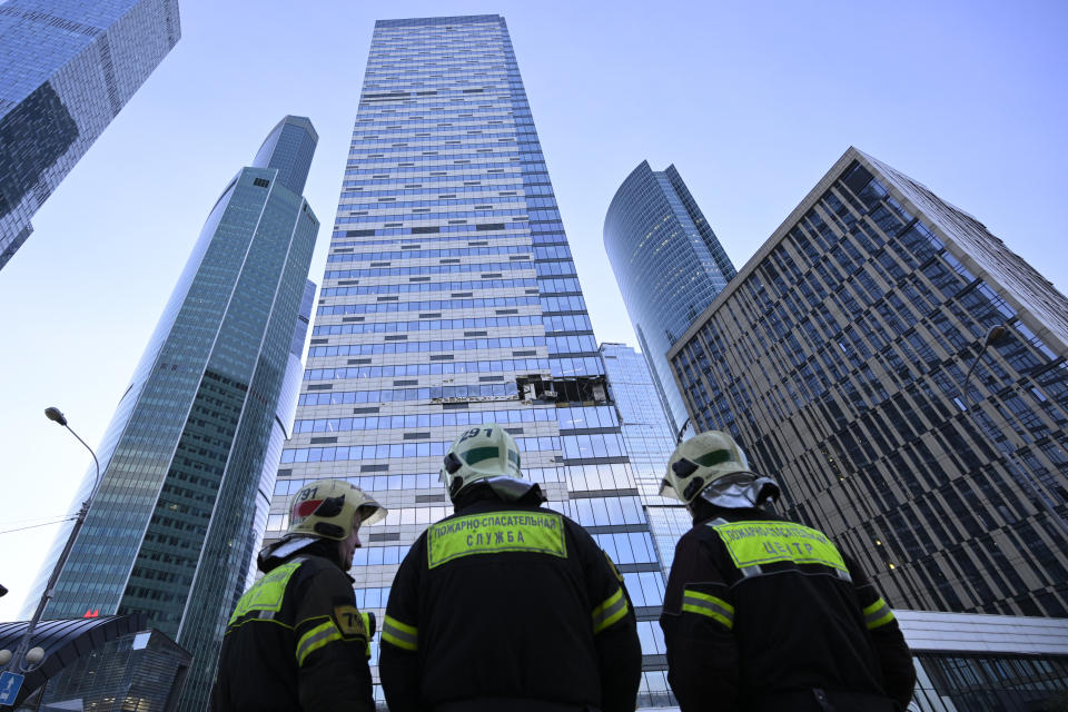 Three workers in hardhats with fluorescent green patches on their work suits look up at the damaged skyscraper.
