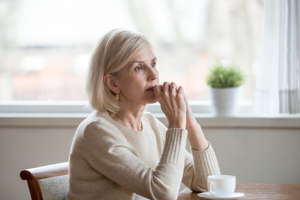 Woman spend time at home alone sitting at table with cup of tea folds hands on chin lost in thoughts. Old lonely female has health problem or thinking about life, reminiscing the past relive memories