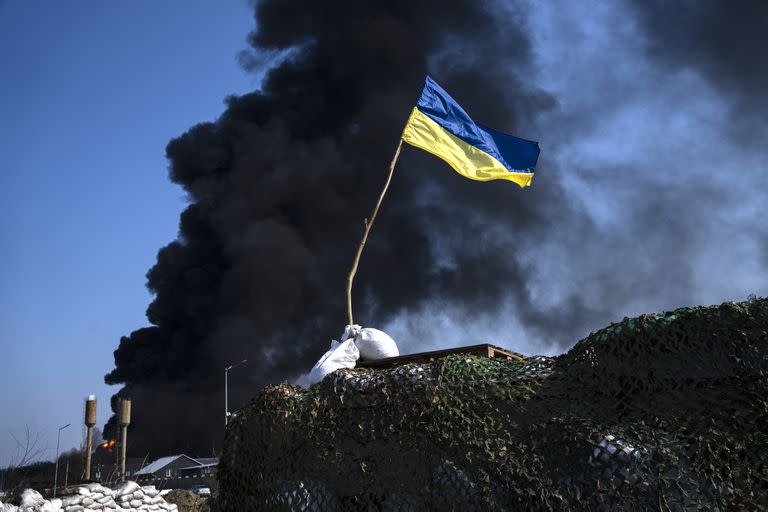 A Ukrainian flag is seen on top of a checkpoint as black smoke rises from a fuel storage of the Ukrainian army following a Russian attack, on the outskirts of Kyiv, Ukraine, Friday, March 25, 2022. (AP Photo/Rodrigo Abd)