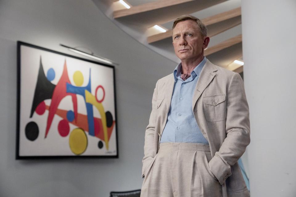 Daniel Craig is standing indoors in a light suit and blue shirt, with a modern abstract painting in the background