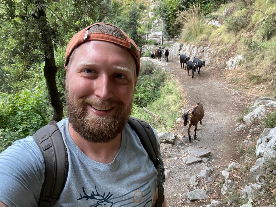 Timothy poses for a selfie as goats follow behind him.