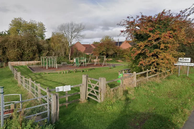 A playground is pictured in a small grassed area with a number of items of play equipment and a gate in the foreground. A sign that says 'Hoby - please drive carefully' is at the edge of the image.