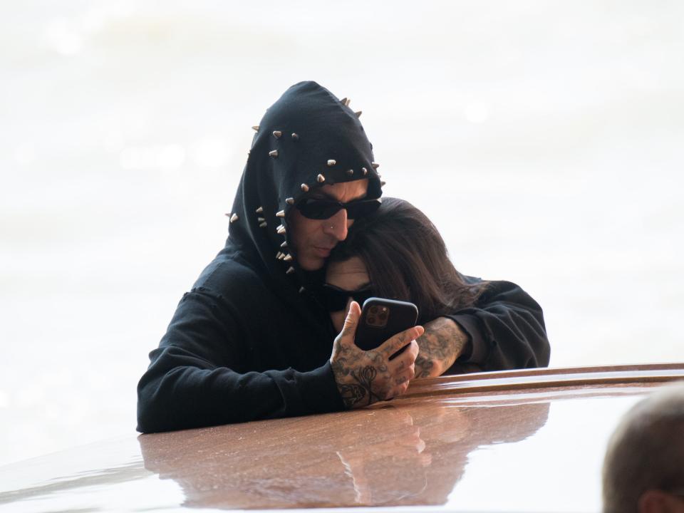 travis barker cuddling kourtney kardashian to his chest as they both look at his cell phone. they're both wearing black clothes and sunglasses