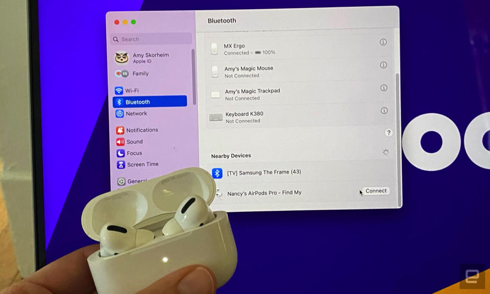 An AirPods case with the white pairing light glowing is held near a MacBook screen with the Bluetooth menu open. 