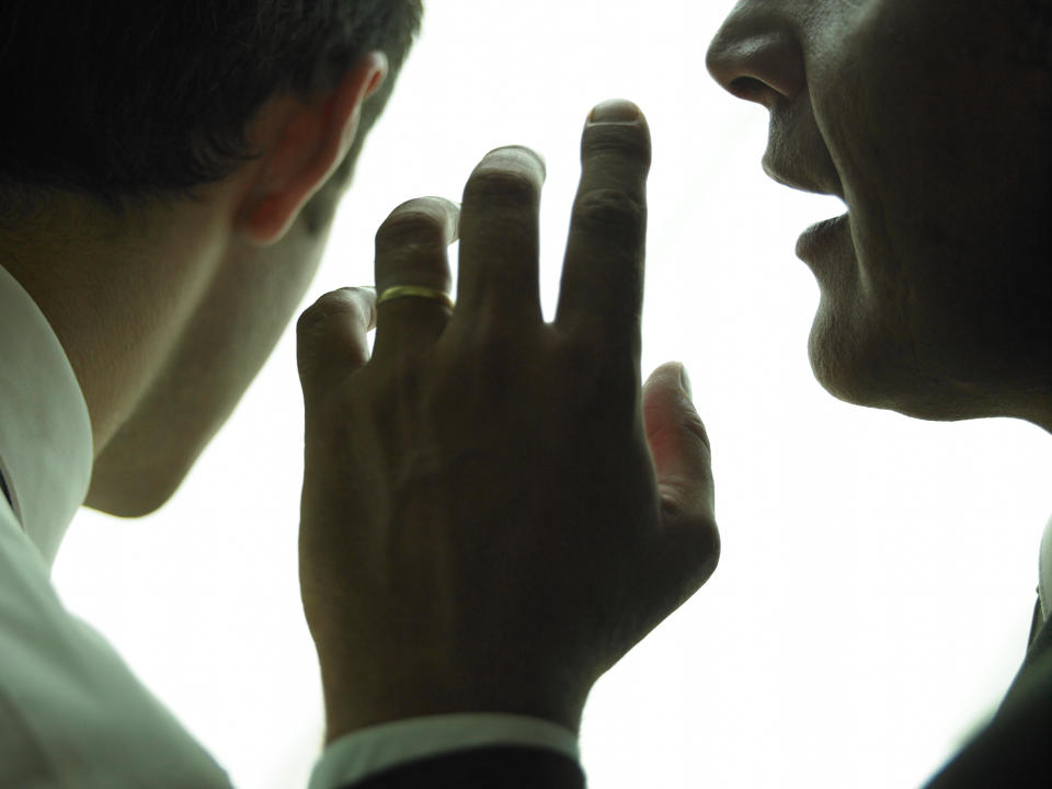 Two people talking closely, one whispering to the other, hand raised to mouth. Faces are not fully visible