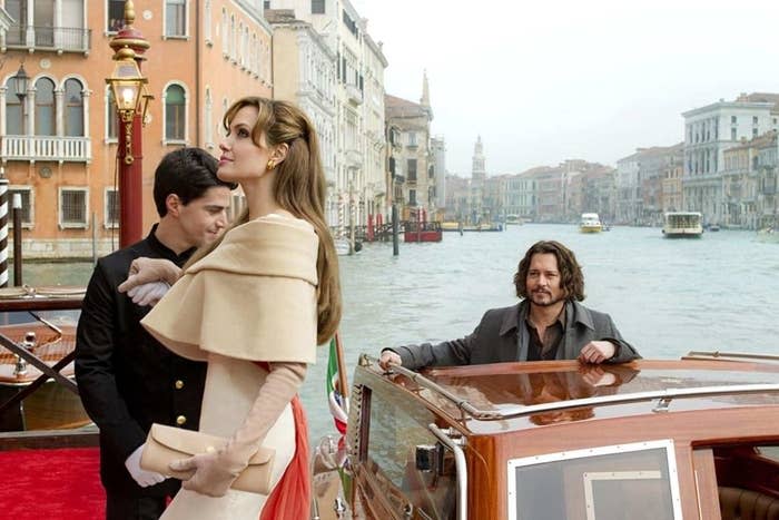 Angelina Jolie in a chic beige cape, planting a kiss on a man's cheek, while another man stands by a boat in Venice