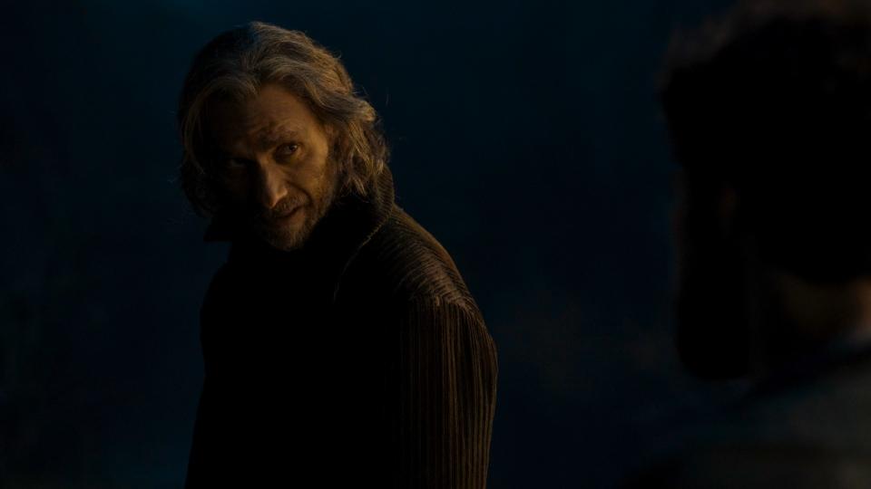 A shot of Thom Merrilin (Alexandre Willaume), the Gleeman, at night. Thom has long, salt-and-pepper hair and a short beard. He wears a brown, textured coat