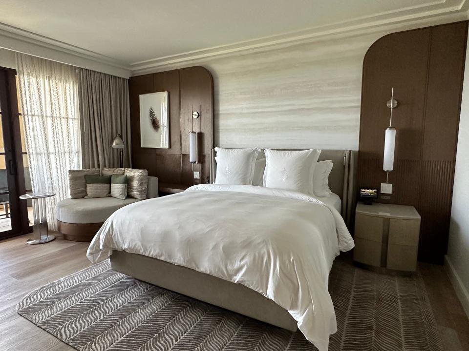 A hotel bed with four pillows and a brown leather headboard. On either side are wood panels with thin cylindrical lights. On the left side is a framed photo of a feather and a gray loveseat with green-and-gray-striped pillows next to a glass side table and a glass door to a balcony, with long panel curtains in front. The carpet under the bed has a wavy brown abstract design.