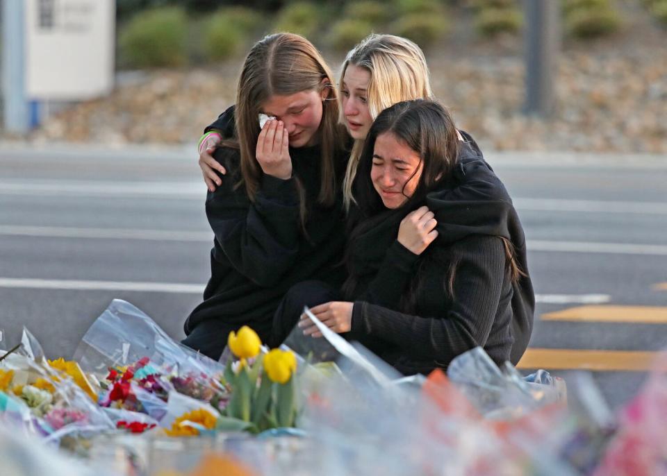 Three teen girls embrace and cry next to a roadside memorial of flowers.