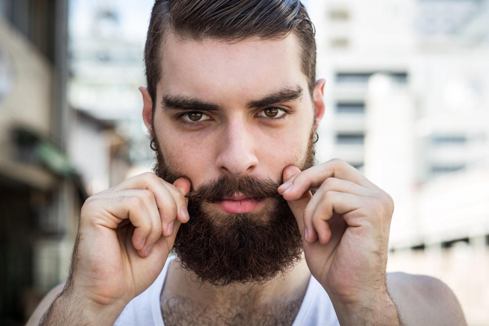 A bearded hipster twists the ends of his mustache whilst looking into the camera