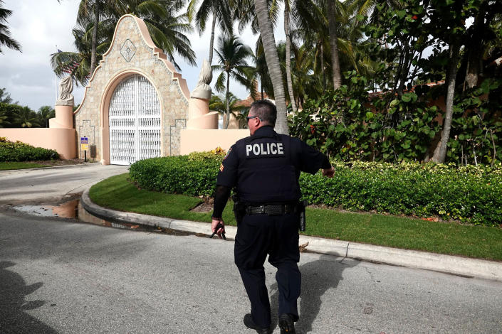 A uniformed police officer is seen from behind while walking along a driveway lined by palm trees.