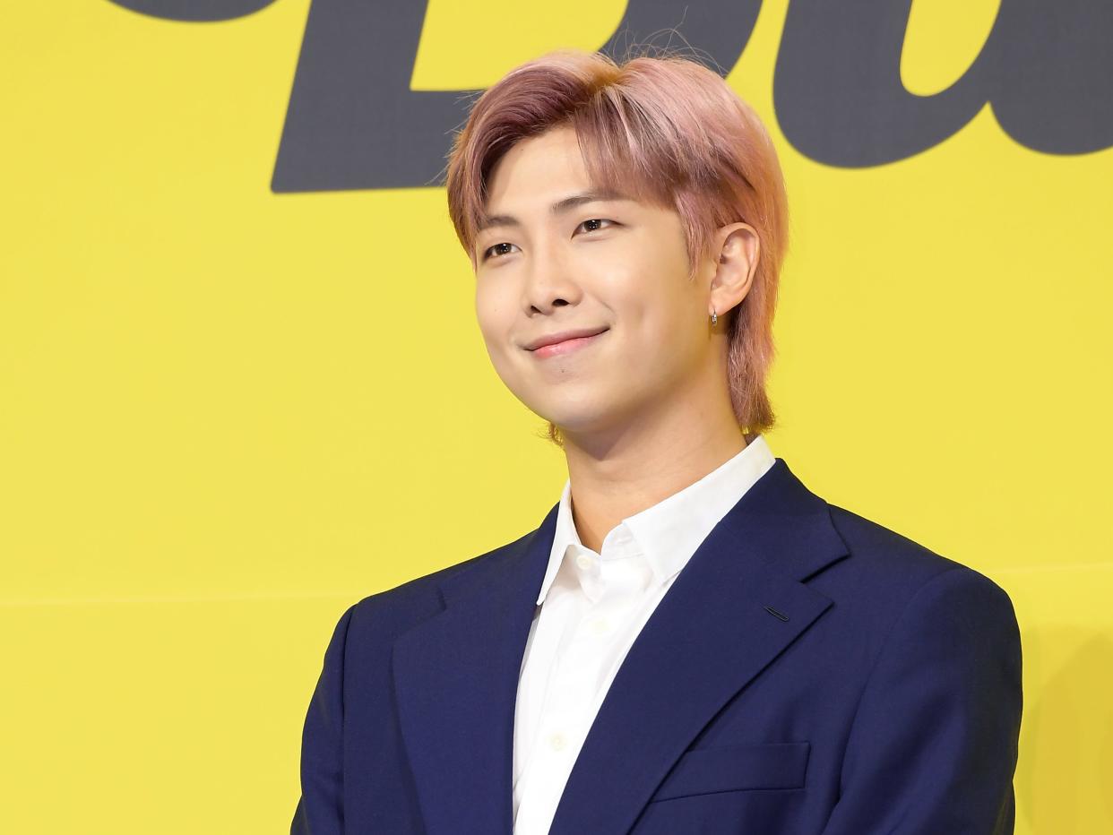 rm from bts standing in front of a yello background. he has pink hair, grown out slightly along his neck and styled up from his face. his hands are clasped in front of his waist, and he's smiling slightly
