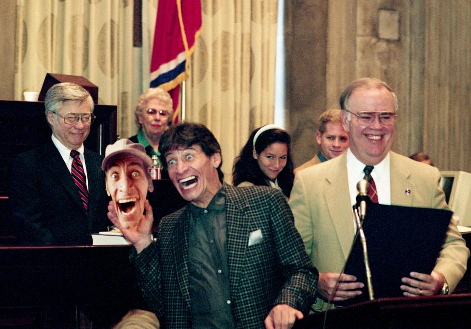 Actor Jim Varney, center, hams it up with a mask of his Ernest character during a ceremony honoring him in the state Senate chamber of the state Capitol March 12, 1992. Chief Clerk Clyde McCullough, right, reads the proclamation honoring Varney.