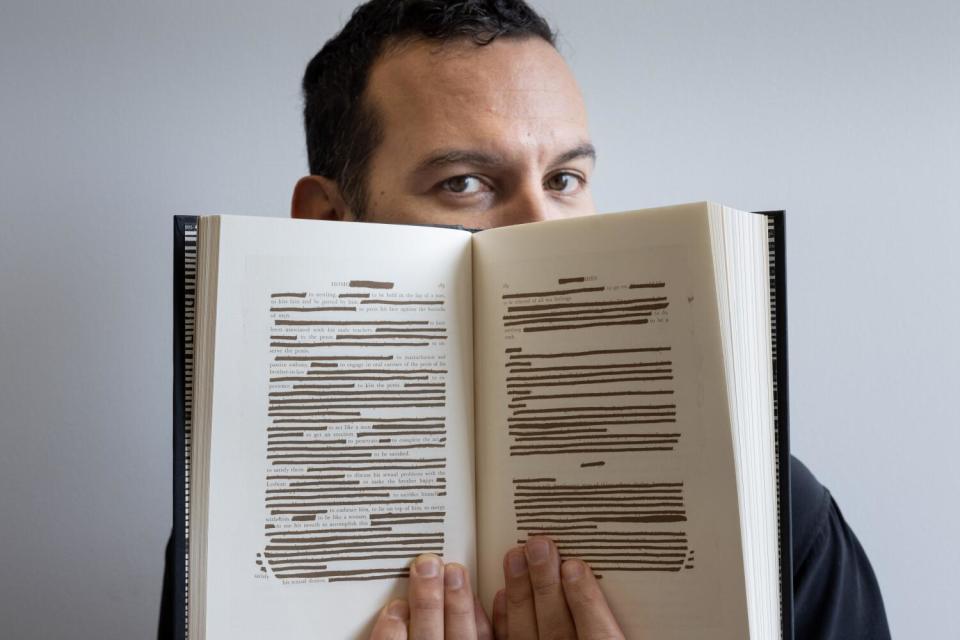 A man holds a book open to pages bearing many words crossed out.