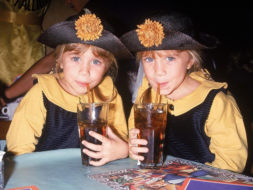 Mary-Kate and Ashley sip from straws in 1992. They wear matching yellow shirts with black jumpers.