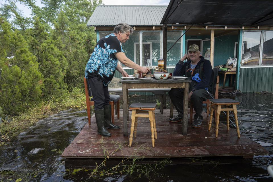 Lyudmila Kulachok, 54, left, sets the food on the table for family diner at the flooded courtyard of her house in the island of Kakhovka reservoir on Dnipro river near Lysohirka, Ukraine, Thursday, May 18, 2023. Damage that has gone unrepaired for months at a Russian-occupied dam is causing dangerously high water levels along a reservoir in southern Ukraine. (AP Photo/Evgeniy Maloletka)