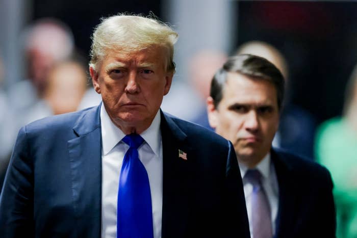 Donald Trump wearing a suit and blue tie, walking followed by another man. Both have serious expressions