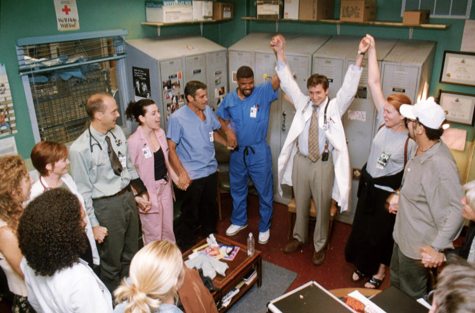 Cast of ER celebrates arm in arm in a crowded medical staff room, including Anthony Edwards, Julianna Margulies, and George Clooney in scrubs and lab coats