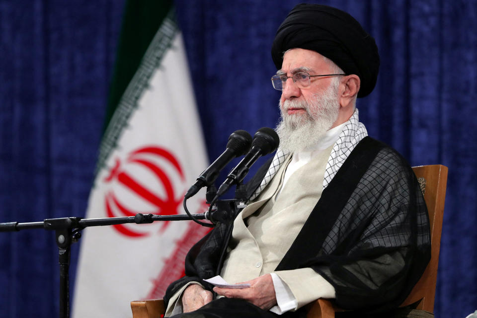 Iran's Supreme Leader Ayatollah Ali Khamenei in black hat and robe, seated at a microphone, with an Iranian flag in the background.