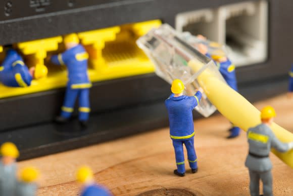 A handful of tiny worker miniatures carry a huge Ethernet plug up to an equally gargantuan network switch, ready to plug it in.