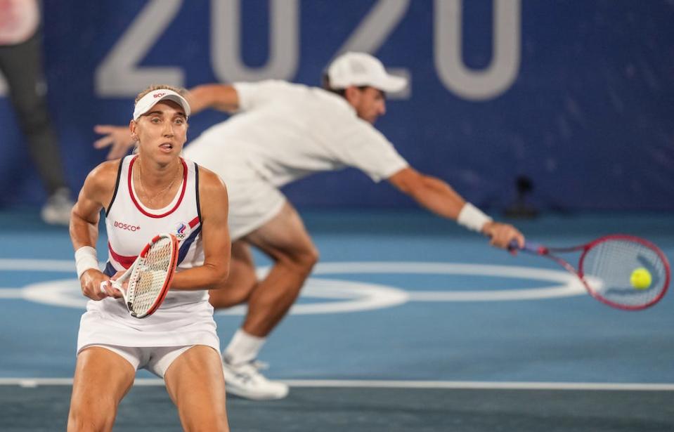 Russian tennis player, Yelena Vesnina stands in readiness at the 2020 Olympic Games mixed doubles tennis tournament with her playing partner, Aslan Karatsev, stretching for the ball in the background, in Tokyo, Japan, August 2021.