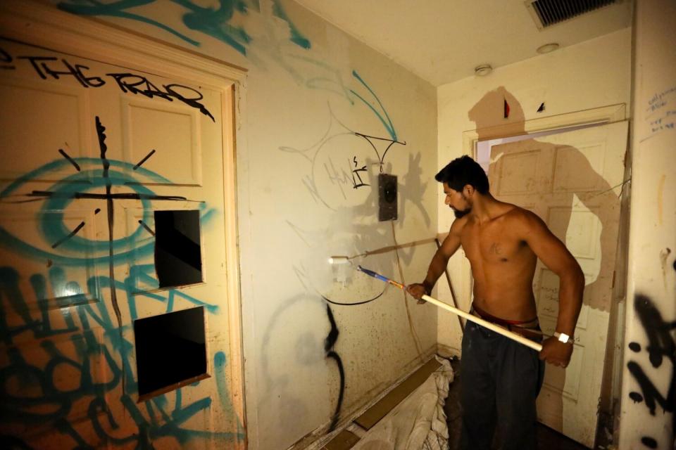 A shirtless man paints over graffiti inside a room