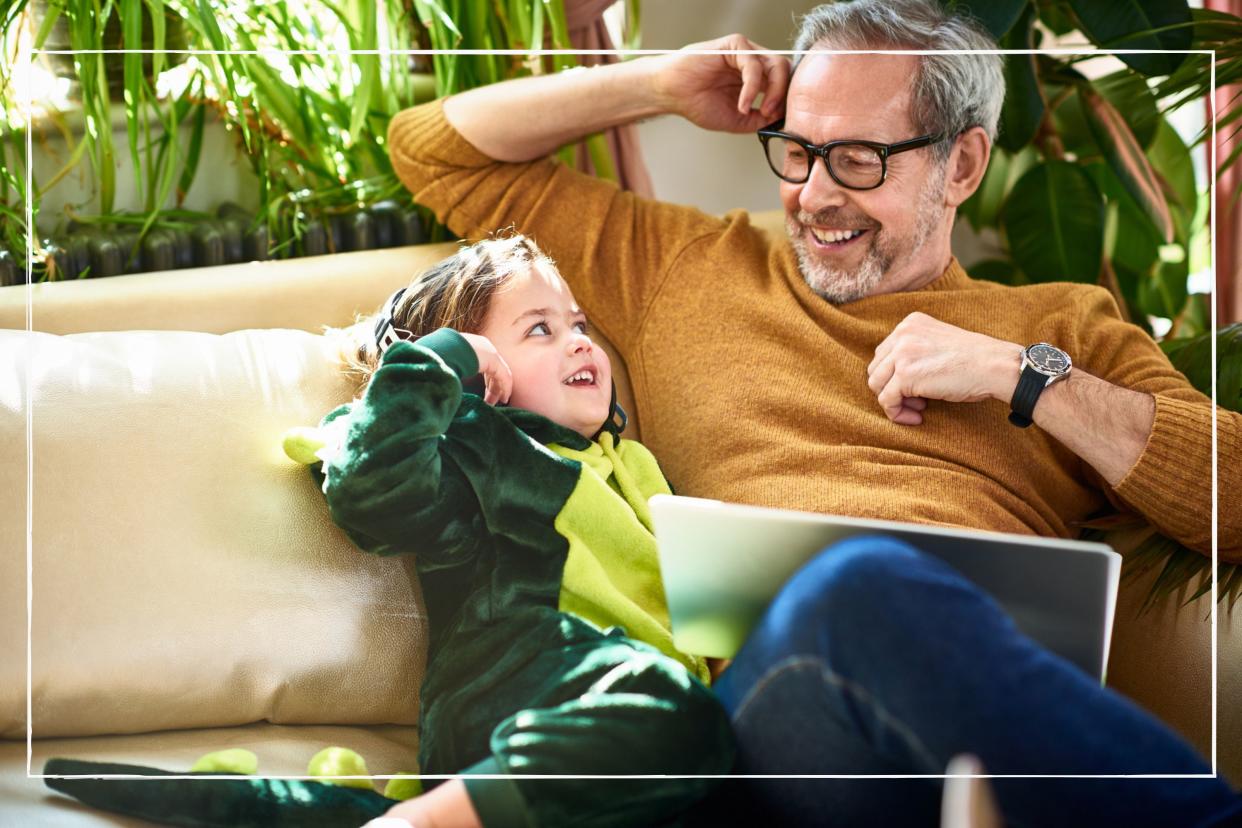  Young grandparent sitting on sofa and smiling with grandchild who is wearing a dinosaur onesie. 