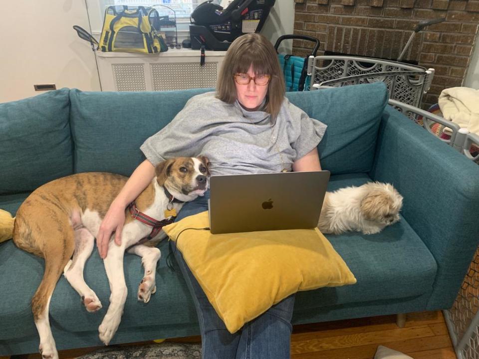 A woman sits on a couch, working on a laptop with a large dog resting beside her and a small dog laying on a pillow