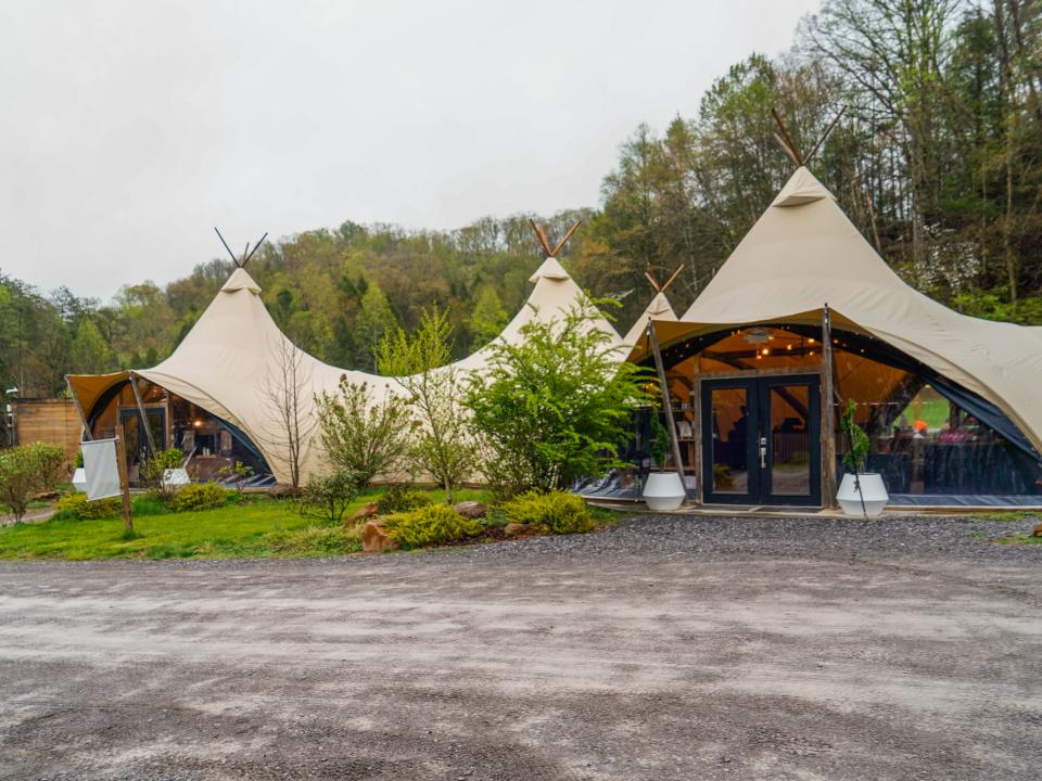 Large four-pointed teepee tents in front of a forest. In the foreground is a gravel road