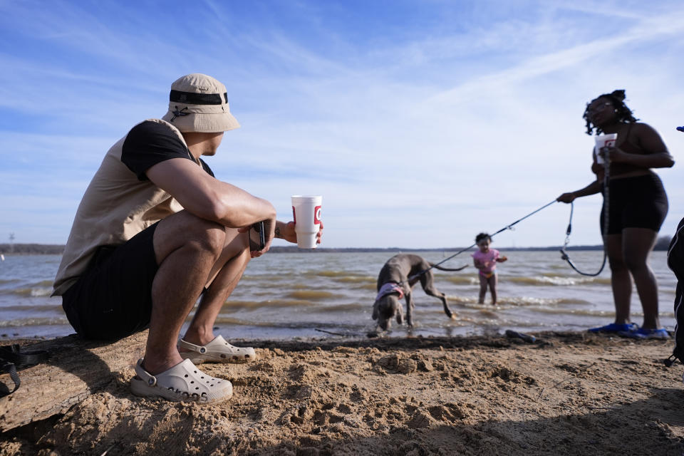 Victor Marcano, left, sits on a log as he watches his daughter Zhamira Marcano, 3, stand in the water on the shore of Joe Pool Lake with Joanna Clarkley, right, and her dog, Pluto, during an unseasonably warm winter day, Monday, Feb. 26, 2024, in Grand Prairie, Texas. (AP Photo/Julio Cortez)