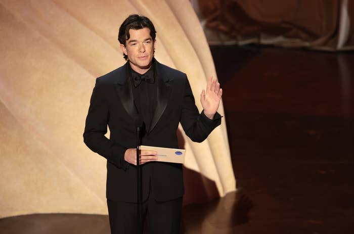 Man in black tuxedo speaking on stage at a celebrity event, holding an envelope