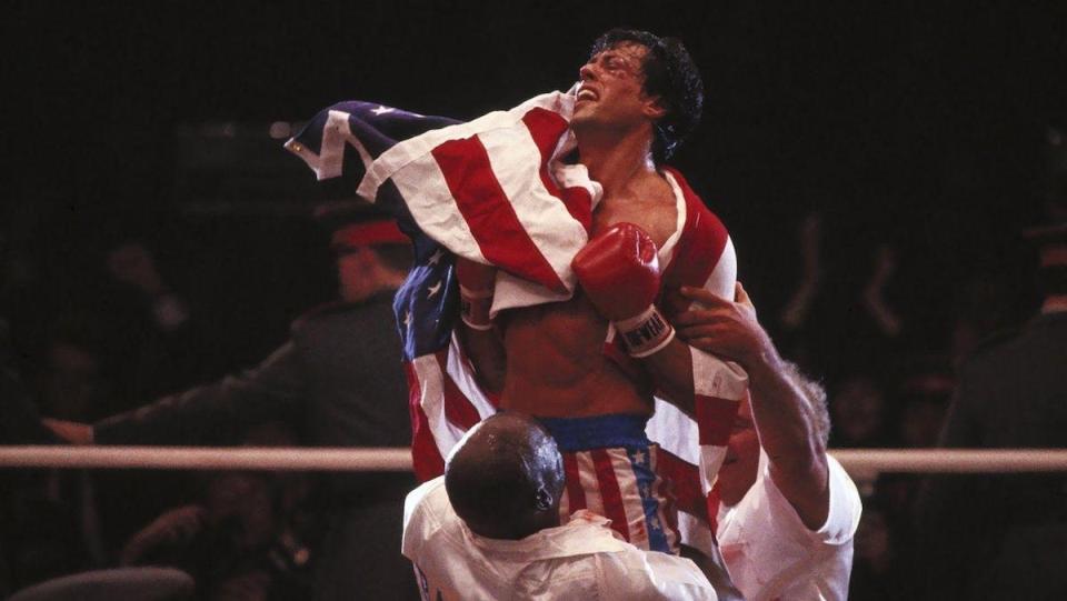 Rocky bloody and battered and draped in the American flag is held up innt he ring after defeating Ivan Drago in Rocky IV