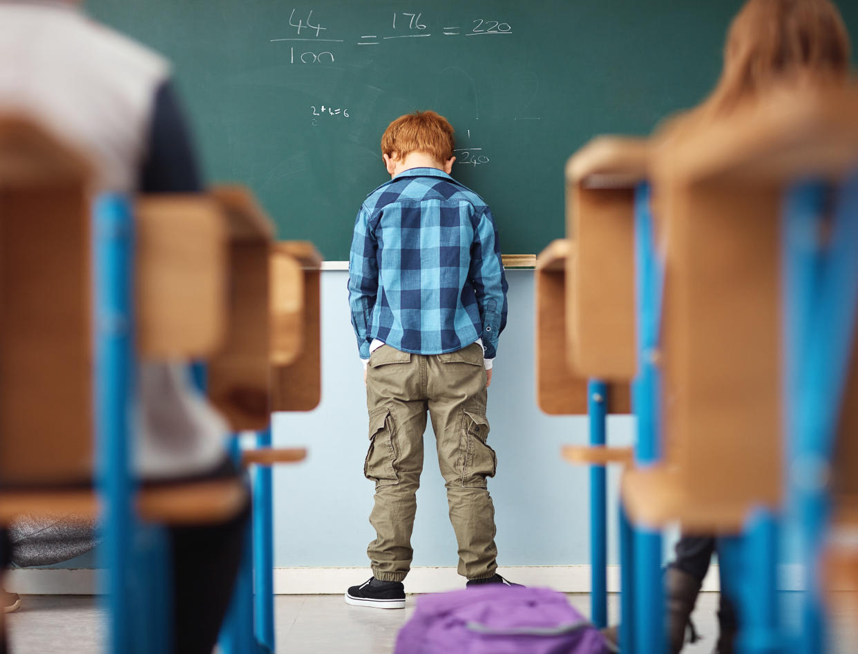 A child stands against a classroom wall, hands in pockets, with his head leaning on a chalkboard.