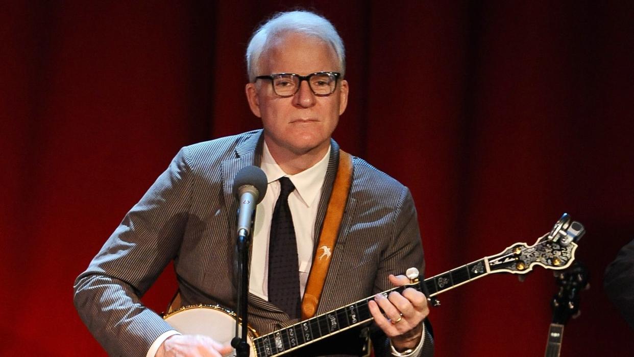 steve martin plays banjo on a stage in front of red curtain, he wears a gray suit with a black tie and white shirt, his glasses are black rimmed