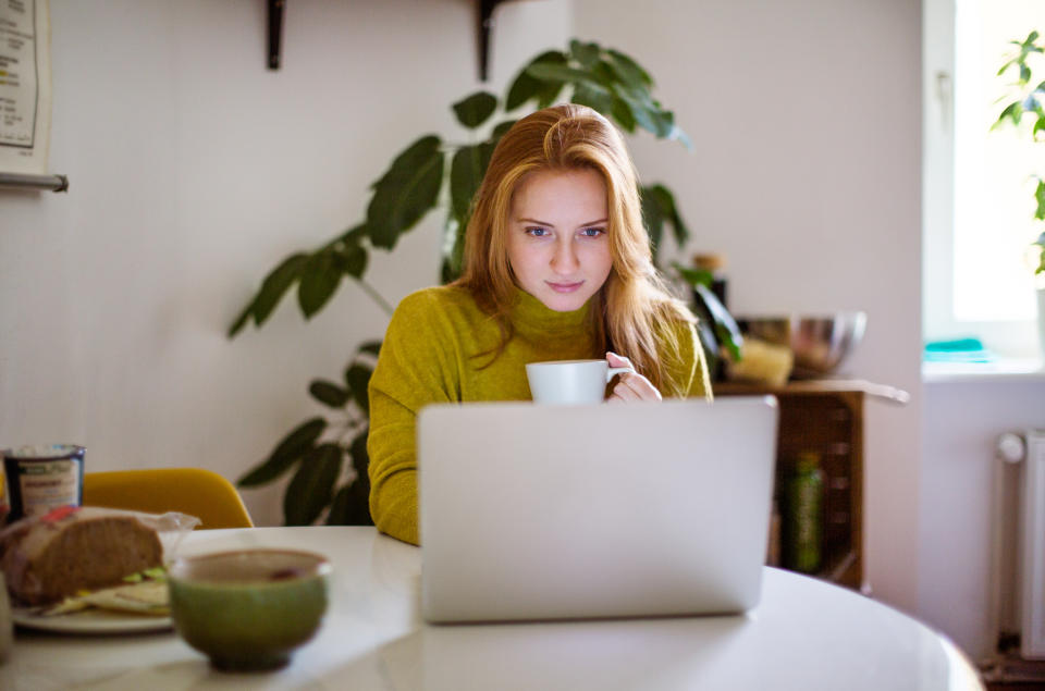 Young woman sitting at table looking at her laptop and drinking coffee. Female using laptop at home.