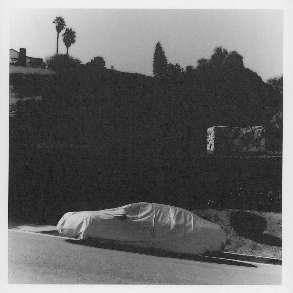 A black-and-white photo of a covered car parked on a sloping street.