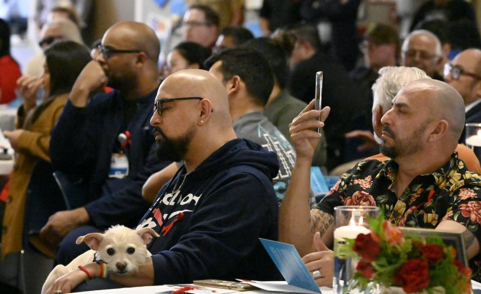 A Black man sits at a table surrounded by a group of other men at a large gathering.