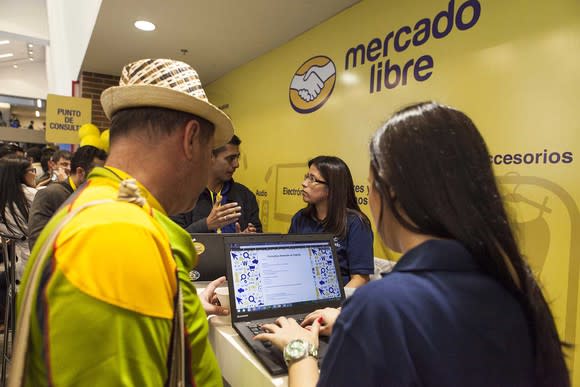 Two people talking in front of a register, with MercadoLibre's logo on a yellow wall behind them.