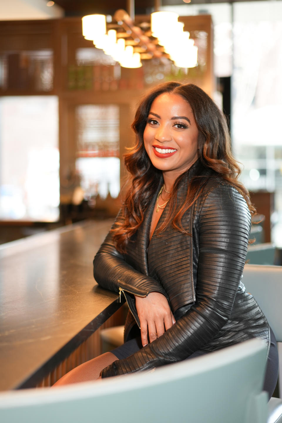 A woman smiles, seated at a bar, wearing a stylish leather dress